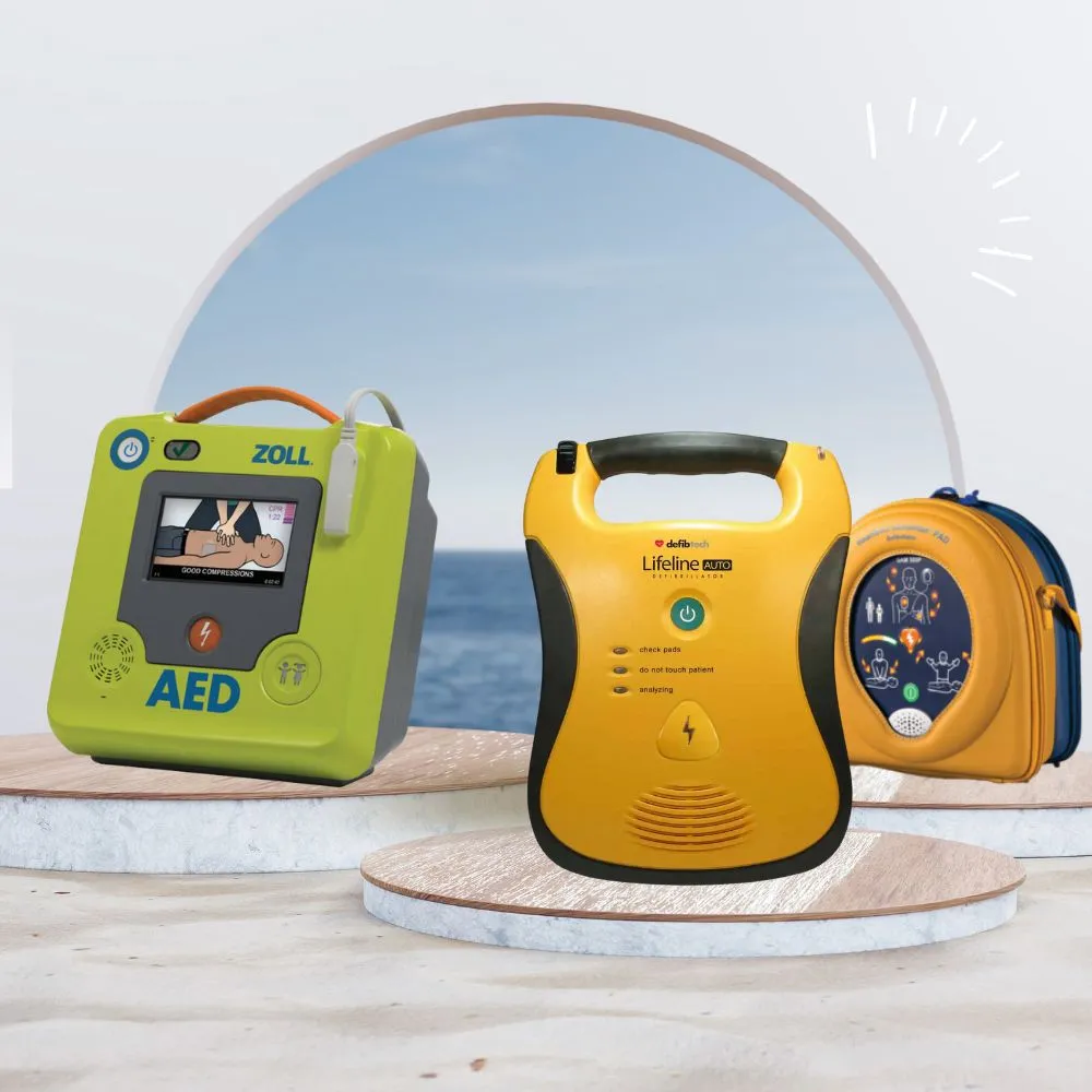The image showcases three different models of automated external defibrillators (AEDs) displayed on circular platforms with a beach and ocean backdrop. From left to right, the models are:  A green and gray AED with the brand name "ZOLL" prominently displayed, featuring a screen showing CPR instructions.
A yellow and black AED with the brand name "Defibtech Lifeline AUTO" and clear labels for operation steps.
An orange and blue AED in a carrying case with the brand name "HeartSine 360P," showing usage instructions on its front panel.
The background features a soft pastel sky with the ocean visible through a large circular frame, creating a serene and inviting atmosphere.