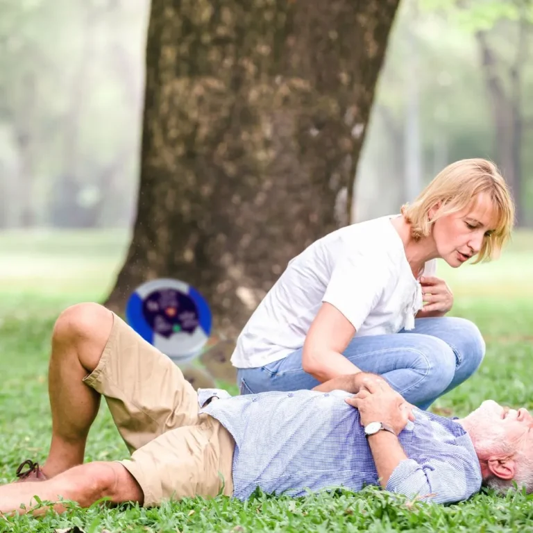 The image shows a woman in a white t-shirt and jeans attending to an elderly man lying on the grass in what appears to be a park. The man is wearing a light blue shirt and beige shorts and seems to be unconscious or in distress. The woman is checking on him, possibly assessing his condition or preparing to perform first aid. The background features trees and a blurred area that looks like part of a park. There is also a defibrillator case or first aid kit partially visible in the background near the tree.