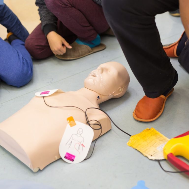 The image shows a CPR training session with a practice mannequin. The mannequin has defibrillator pads attached, and there is a defibrillator visible. The setup is being used to demonstrate or practice the application of an Automated External Defibrillator (AED) in a training environment.